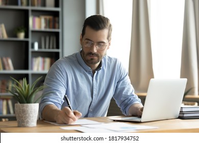 Concentrated Young Caucasian Man In Glasses Sit At Desk Work On Computer Make Notes. Focused Serious Male Worker Or Freelancer Busy At Home Office Watch Webinar On Laptop, Handwrite On Paper.