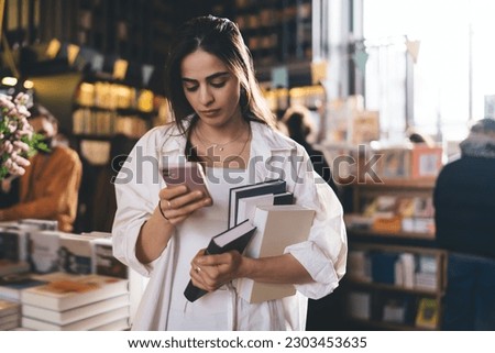 Concentrated young ethnic female in casual clothes standing with books in bookstore while messaging on smartphone during shopping at daytime
