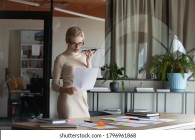 Concentrated young female blonde ceo manager using virtual assistant on phone, checking information in financial paper document, 30s businesswoman leader sending voice mail for client in modern office - Powered by Shutterstock