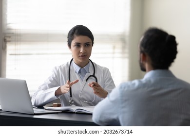 Concentrated young female indian doctor physician giving healthcare consultation to african american male patient, explaining medical insurance benefits or illness treatment at checkup meeting.