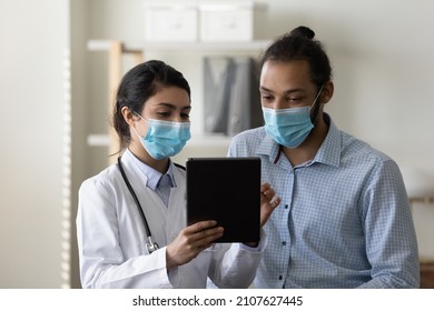 Concentrated Young Female Indian Doctor And African American Male Patient In Protective Facial Mask Looking At Digital Computer Touchpad Gadget Screen, Discussing Health Tests Lab Results In Clinic.