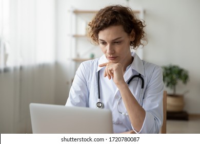 Concentrated Young Female Physician Working On Computer Looking At Laptop Screen Thinking On Problem Solution Concept. Serious Woman Doctor Wearing White Coat Doing Online Research At Workplace.