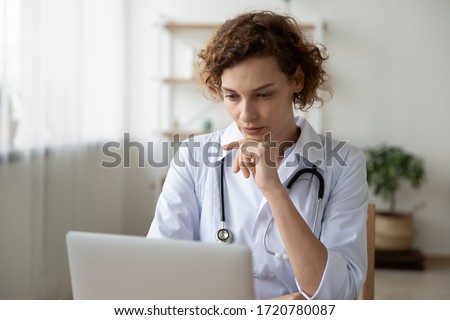 Concentrated young female physician working on computer looking at laptop screen thinking on problem solution concept. Serious woman doctor wearing white coat doing online research at workplace.