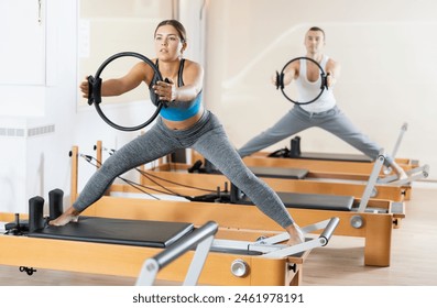 Concentrated young fit girl doing stretching exercises on Pilates reformer, using resistance ring to muscle strength. Concept of pilates as physical therapy alternative for injury rehab.. - Powered by Shutterstock