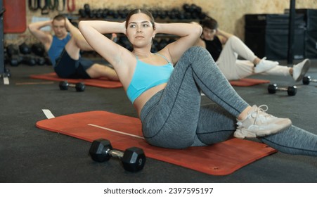 Concentrated young fitness woman doing abdominal exercise on yoga mat during group workout in gym. CrossFit healthy concept.