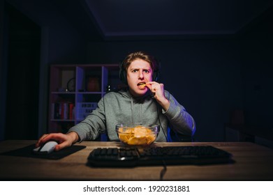 Concentrated young gamer playing online games at night on computer and eating chips with serious face looking at camera. Guy has dinner at night at the computer and streaming games