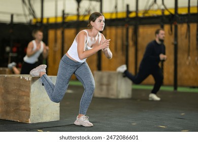 Concentrated young girl in fitness attire performing Bulgarian split squats using wooden plyometric box at gym. Bodyweight training concept - Powered by Shutterstock