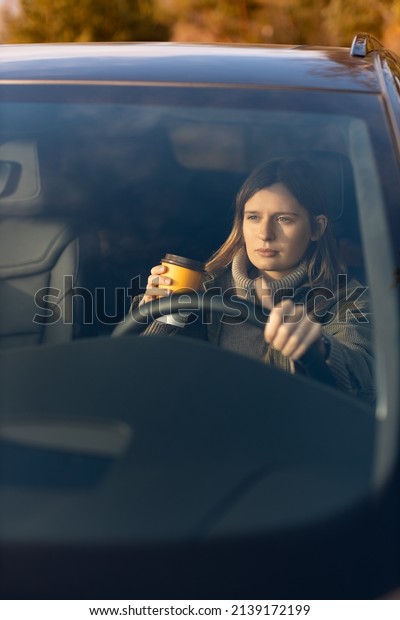 Concentrated young girl in the morning goes to\
work by car and drinking coffee. View through the windshield. Copy\
space. Driving car\
concept.