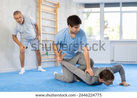 Concentrated young guy practicing armlock, painful control move to hold and immobilize opponent, in training bout with male partner during self defence course in gym