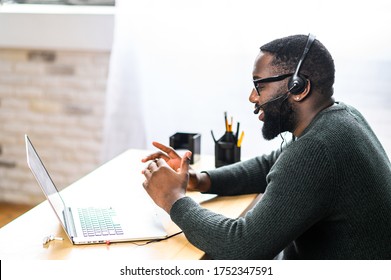 Concentrated young guy in smart casual wear and stylish glasses is using headset and laptop for online communication, supporting, selling. A black guy sits at the office desk, side view