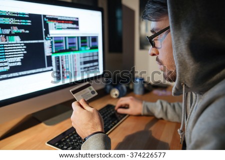 Concentrated young hacker in glasses stealing money from different credit cards sitting in dark room