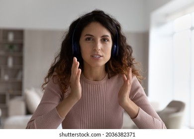 Concentrated young hispanic woman in headphones looking at camera involved in distant web camera meeting, communicating distantly by video call, virtual event, online conference vlogging concept.