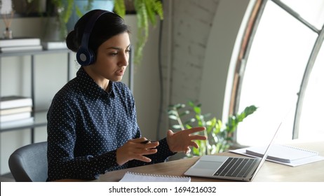 Concentrated young indian businesswoman wearing wireless headset, holding educational video conference call with partners, discussing project details with client online, working remotely from home.