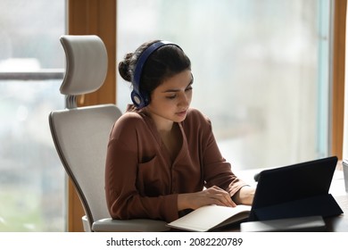 Concentrated young indian ethnicity business woman in wireless headphones holding distant video call meeting using digital computer tablet, writing notes in paper planner or studying on online courses