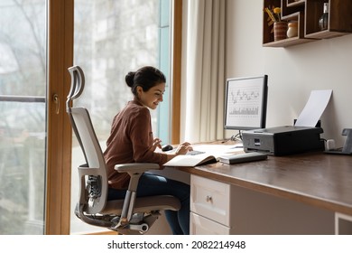 Konzentrierte junge indianische Ethnizität Frau sitzt in komfortablen, verstellbaren ergonomischen Sesseln mit Lumbalstütze, studiert oder arbeitet in einem modernen Heimbüro am Computer. Fernarbeitskonzept.