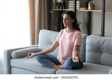 Concentrated Young Indian Female Sitting On Sofa With Crossed Legs Practicing Yoga Pranayama Exercises. Calm Relaxed Millennial Arabic Woman Meditating At Home Taking Lotus Position Relieving Stress