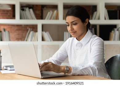 Concentrated young Indian female worker sit at desk look at laptop screen consult client or customer online. Mixed race ethnicity woman employee type text on computer in office. Technology concept.