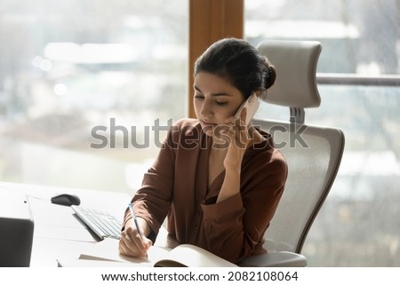 Concentrated young indian woman working in modern home office, holding phone call conversation writing notes in paper planner, giving distant cellphone consultation to client or talking with client.