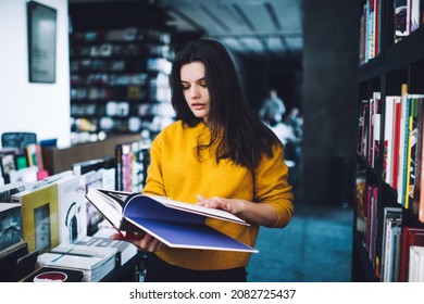 Concentrated young lady in casual clothes reading interesting book in hardcover while standing between bookshelves in contemporary bookshop during daytime