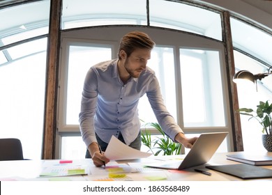 Concentrated Young Male Project Manager Software Developer Standing By Desk At Workplace In Office Involved In Paperwork, Checking Comparing Data In Hardcopy And On Laptop Screen, Making Notes Changes