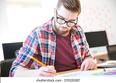 Concentrated young man with beard in plaid shirt and glasses making sketches with pencil sitting at the table - Powered by Shutterstock