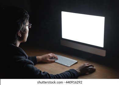 Concentrated young man in glasses sitting at the table and using computer in dark room