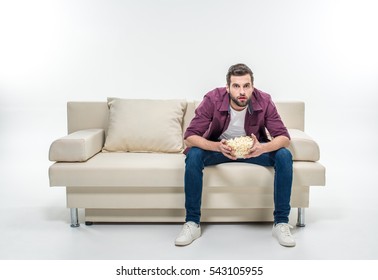 Concentrated Young Man Sitting On Couch With Popcorn Isolated On White