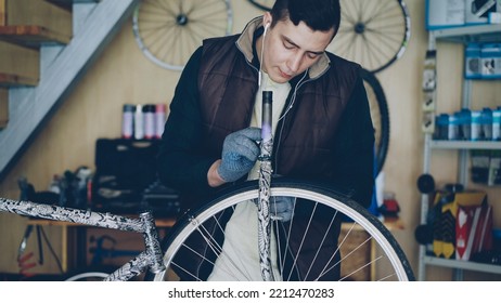 Concentrated Young Mechanic Is Greasing Bicycle Wheel And Listening To His Favourite Music With Earphones While Repairing Bike In His Small Home Studio. Maintenance And People Concept.