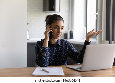 Concentrated young mixed race businesswoman discussing project with partner by phone looking at pc screen. Focused indian female freelancer holding cell at ear using laptop to get information online