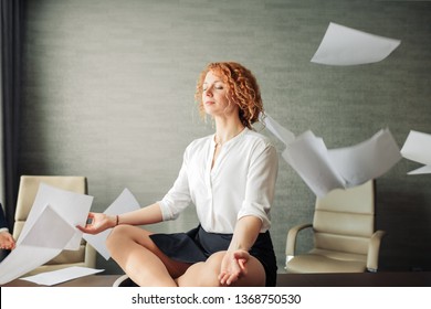 Concentrated young red-haired woman in office clothes sitting on table in lotus position with closed eyes, meditating despite of flying papers chaos surrounding her at office