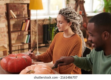 Concentrated Young Woman With Dreads Sitting At Table With Black Guy And Cutting Into Pumpkins While Making Jack-o-lantern For Halloween