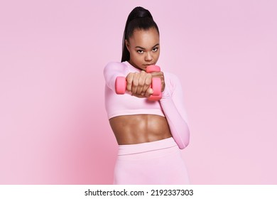 Concentrated young woman exercising with dumbbells while standing against pink background - Powered by Shutterstock