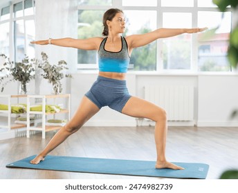 Concentrated young woman performing one-legged balancing asana Virabhadrasana, or Warrior pose during daily yoga routine in cozy airy fitness studio.. - Powered by Shutterstock