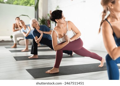 Concentrated young woman performing Revolved Side Angle Pose Parivrtta Parsvakonasana during yoga class with female group in fitness studio - Powered by Shutterstock