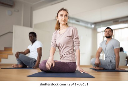 Concentrated young woman performing seated twisting asana Parivrtta Ardha Padmasana during group yoga class in fitness studio.. - Powered by Shutterstock