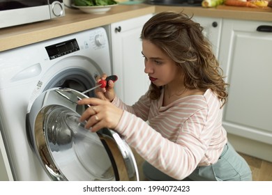 Concentrated Young Woman With Screwdriver Repairing Broken Washing Machine. Young Female Fixing Washer. Breakdown And Repair Of Household Appliances. Kitchen Interior On Blurred Background