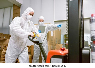 Concentrated Young Worker Wearing Coverall And Safety Mask Using Hand Pallet Truck While Working In Warehouse Of Pharmaceutical Factory, His Superior Using Digital Tablet And Guiding His Actions