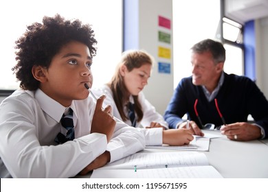 Concentrating Male High School Student Wearing Uniform Working At Table With Teacher Talking To Pupils In Background