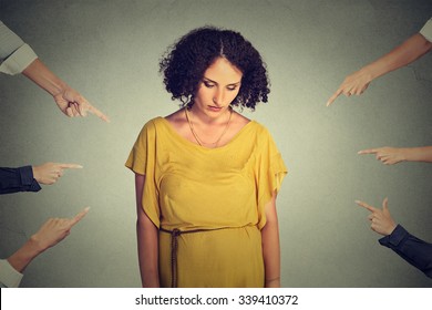 Concept of accusation guilty person girl. Sad embarrassed upset woman looking down many fingers pointing at her back isolated on grey office wall background. Human face expression emotion feeling