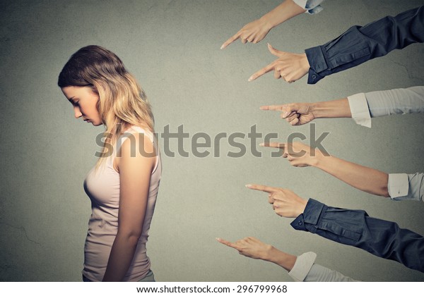 Concept of accusation guilty person girl. Side\
profile sad upset woman looking down many fingers pointing at her\
back isolated on grey office wall background. Human face expression\
emotion feeling