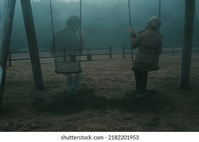 Concept of afterlife and memory for parents or lost love. Back view of woman sitting on a swing with ghost man near her outdoor in the park with fog. . Dead friend or husband concept. Life and death - Shutterstock ID 2182201953