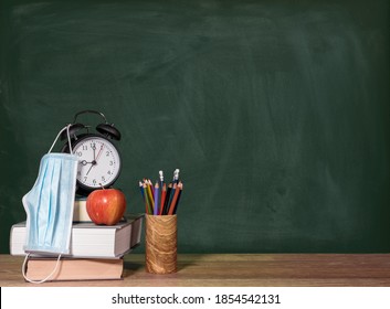 Concept For Back To School With Coronavirus Or Covid-19 With Books, Apple And Alarm Clock Against A Chalkboard With Face Mask