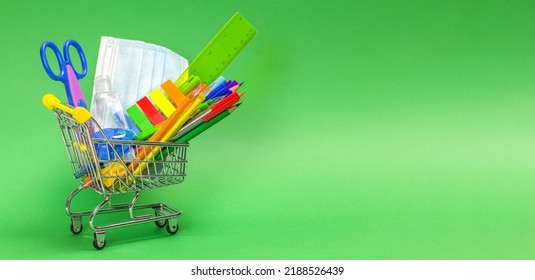 The Concept Of Back To School During The Epidemic. School Supplies And Stationery In Bright Colors, A Medical Mask And A Sanitizer Are In A Mini-cart On A Green Background. Shopping