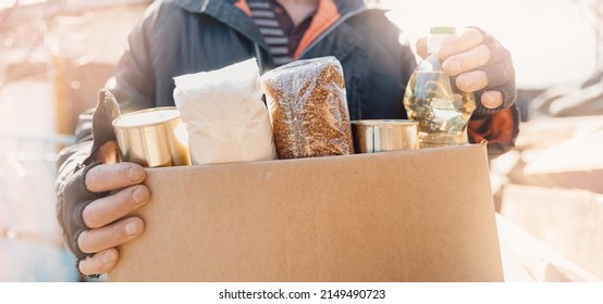 Concept Banner Humanitarian Aid To Refugees. Old Man Senior Is Holding Cardboard Box Of Donations Canned Food, Looking Camera.