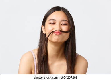 Concept of beauty, fashion and makeup products advertisement. Close-up portrait of happy silly asian girl making fake moustache out of hair strand, smiling cheerful, white background - Powered by Shutterstock