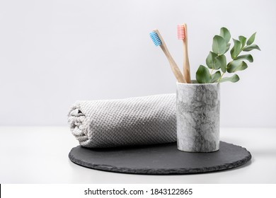Concept of biodegradable objects. Two bamboo toothbrush in cup near bathroom towel and eucalyptus plant on black plate stand against white copy space background. Oral and dental care