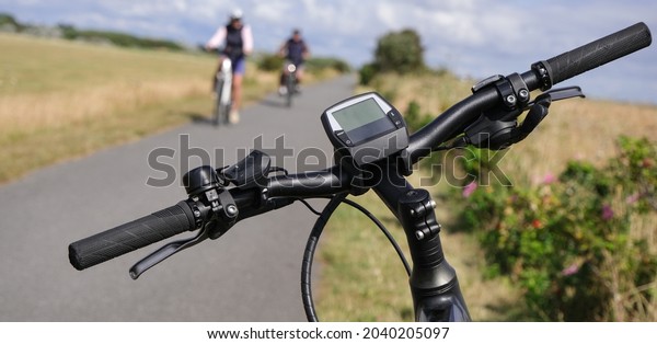 Concept: break at a bike path.
Close-up of a handlebar of an e-bike in the foreground, cyclists,
path and nature in the background, landscape format, selective
focus