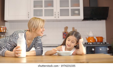 The concept of breakfast in the kitchen. Young mother pours milk on the plate of her cute little daughter, having breakfast with cereal. He begins to eat with pleasure.