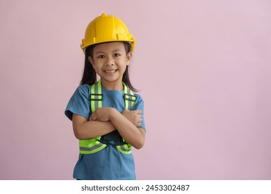 Concept of building, construction, and professions. A little girl in a helmet and safety vest on a pink background smiles standing with her arms crossed. Engineering concepts, energy, design. - Powered by Shutterstock