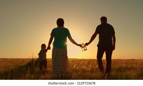 Concept Of Building Family House. Real Estate Investment. Family Holds Paper House In Their Hands, Child Walks With His Parents At Sunset. Dream About Buying House, Happiness, Peace, Family, Home